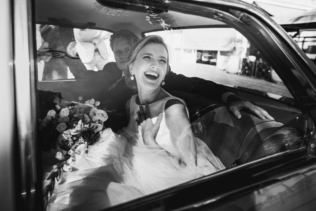 La mariée rie aux éclats au moment de quitter les lieux de sa cérémonie religieuse en voiture.